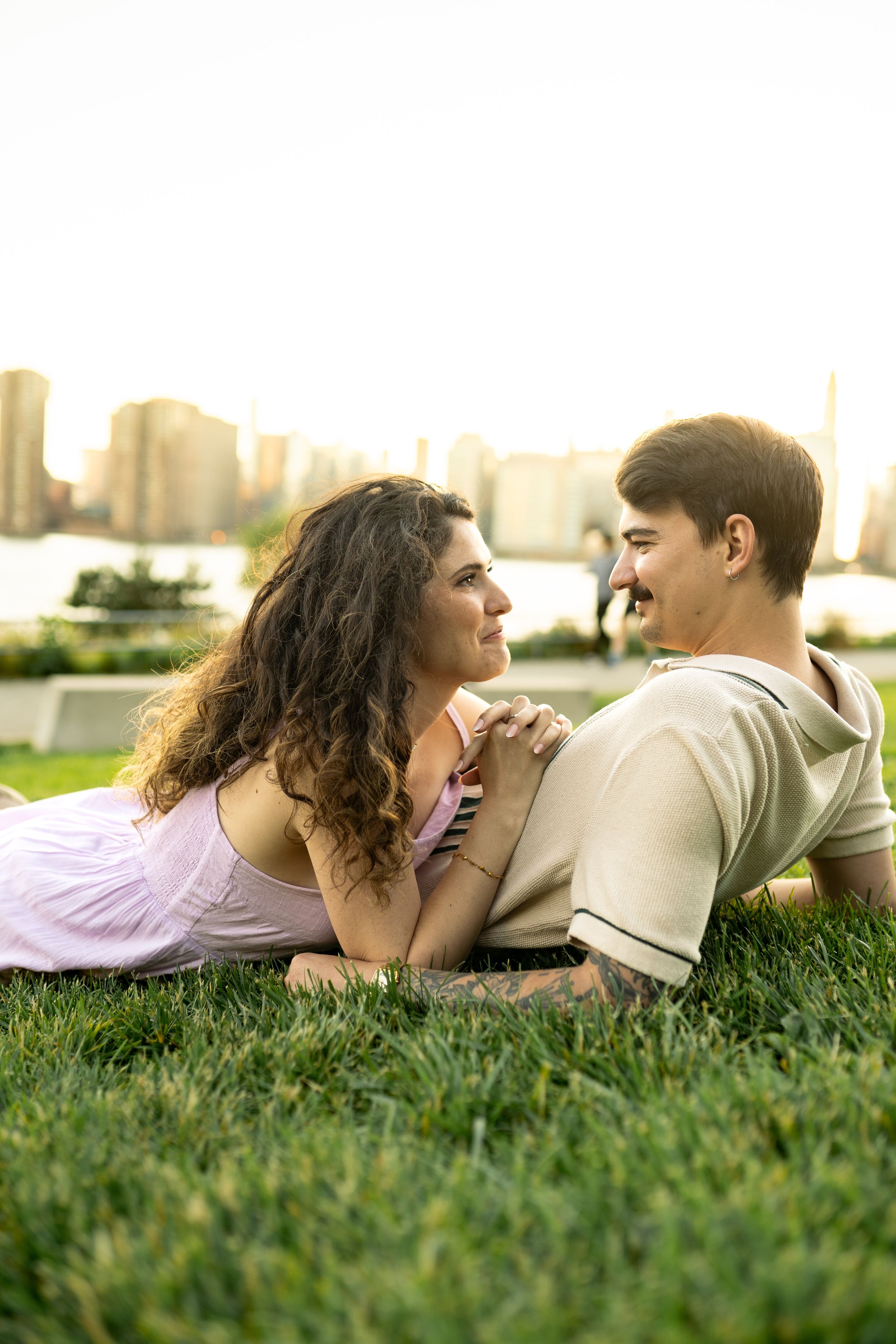 A couple lies together on grass overlooking a waterfront cityscape at sunset.