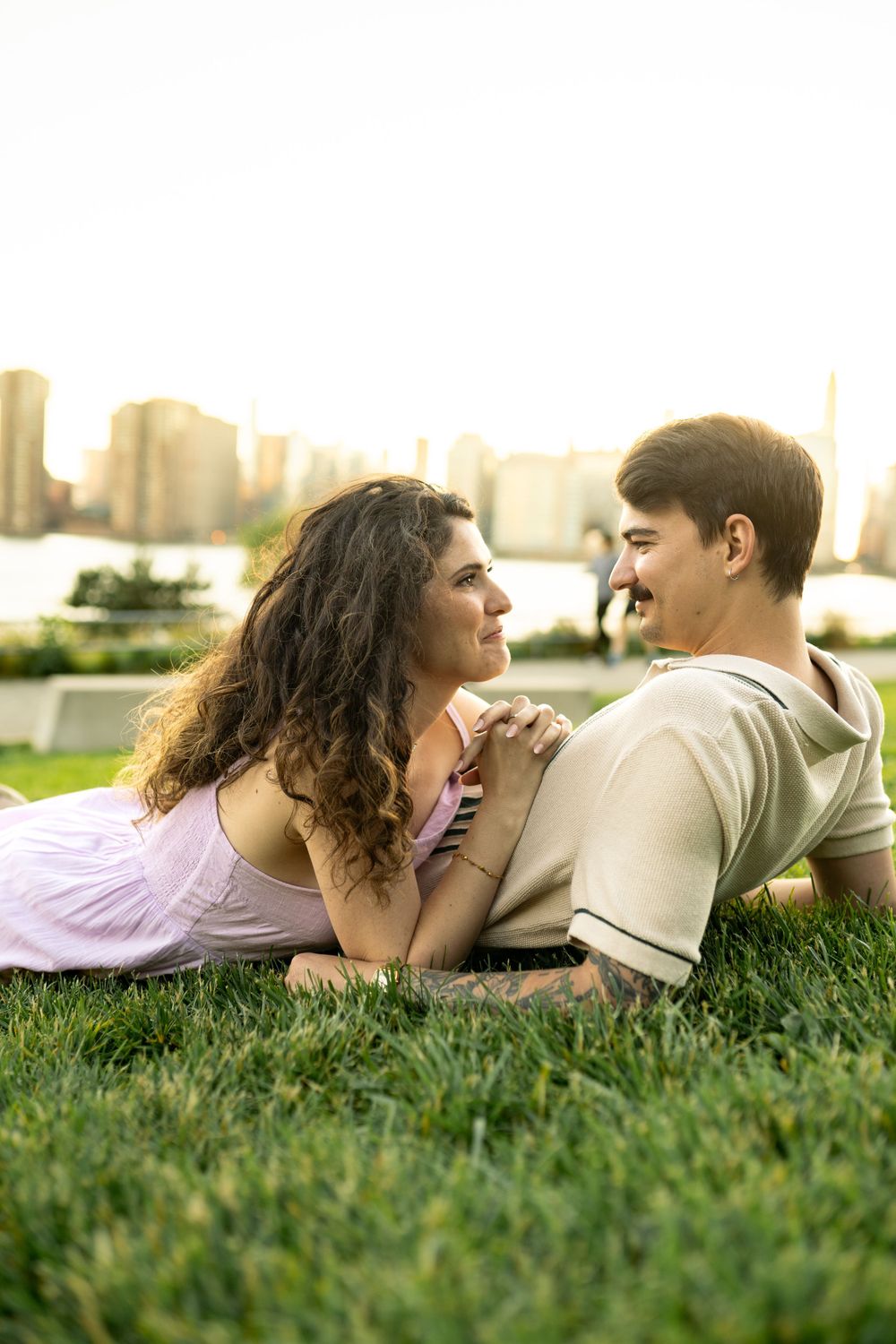 A couple lies together on grass overlooking a waterfront cityscape at sunset.