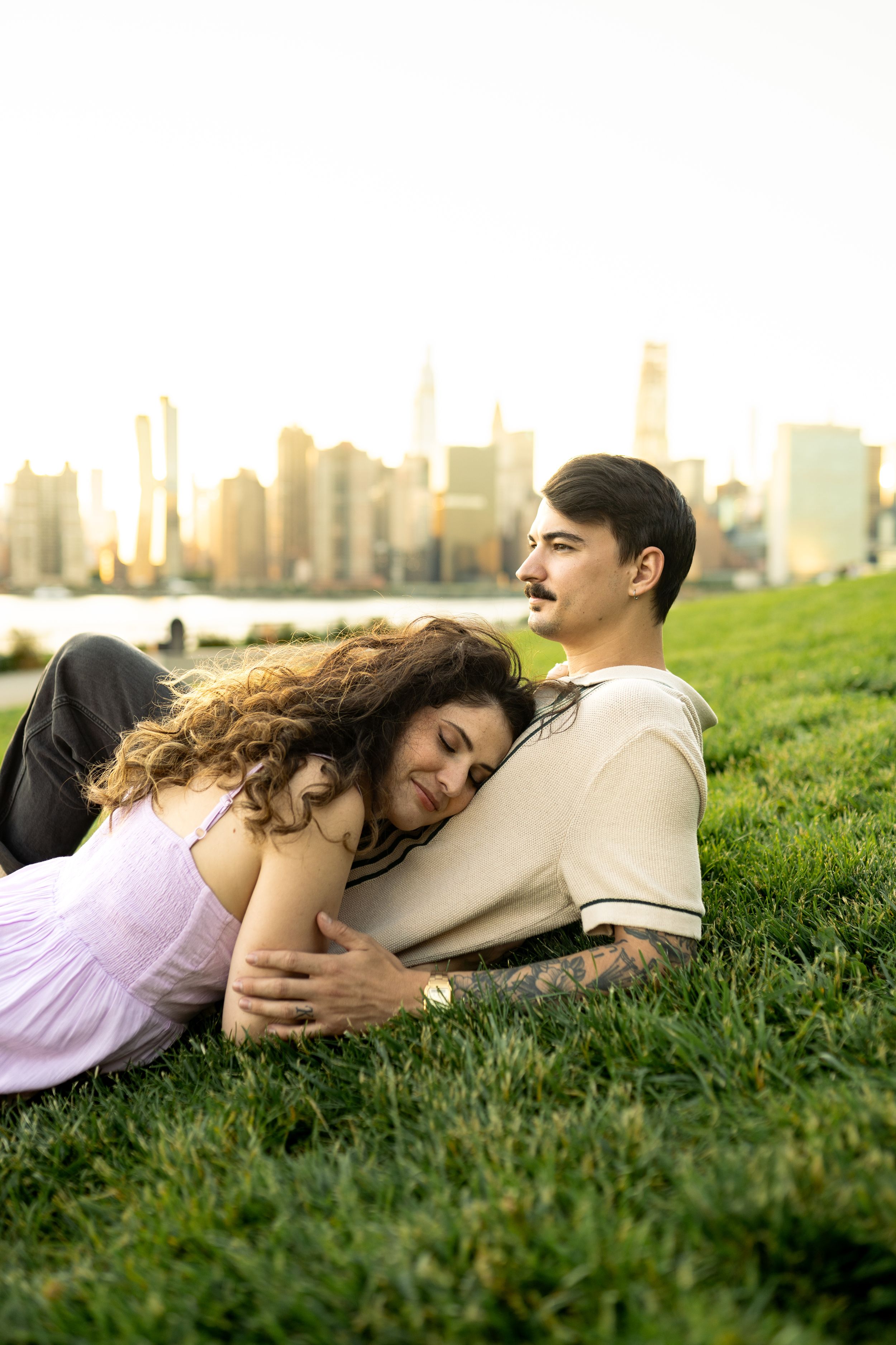 A couple relaxes on grass with the Manhattan skyline visible in the background during sunset.