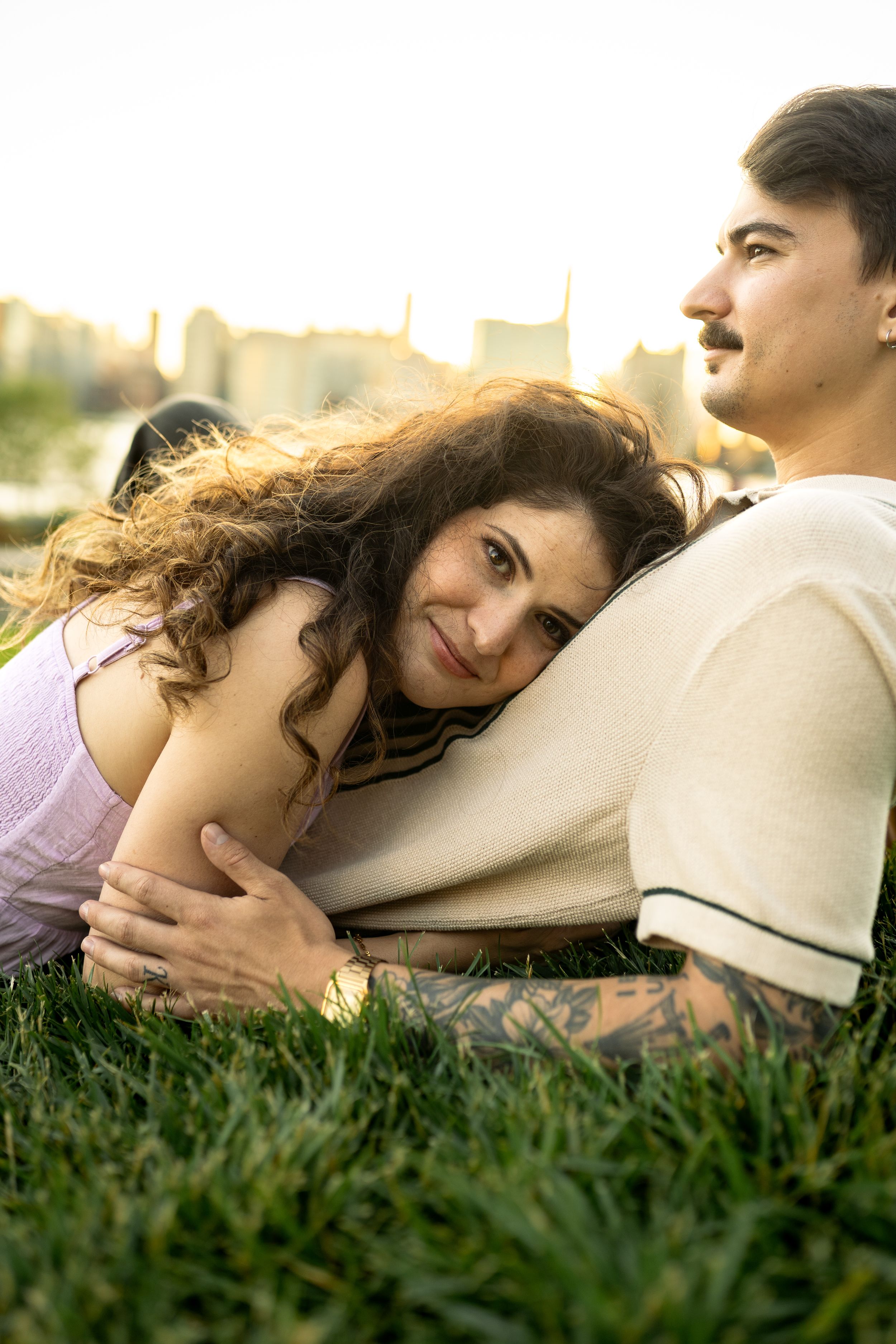 A couple shares an intimate moment while lying on grass with the city skyline behind them.