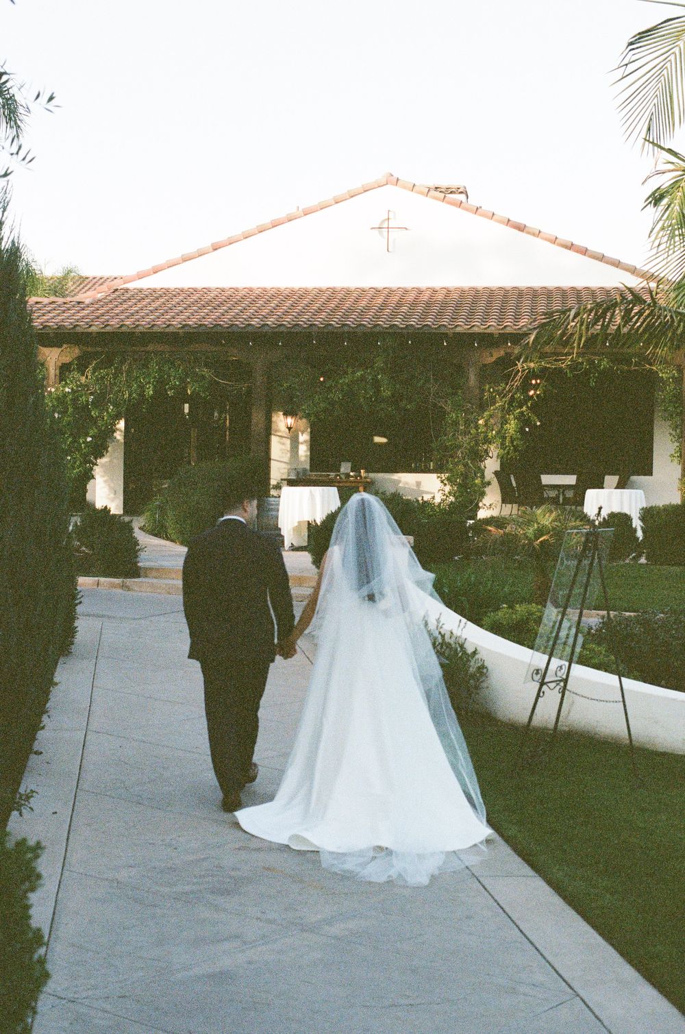 Elegant wedding processional on a stone pathway beside a Spanish mission-style building.