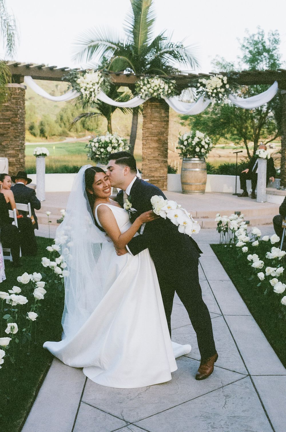 Wedding couple dancing on an outdoor patio decorated with white flowers and palm trees.