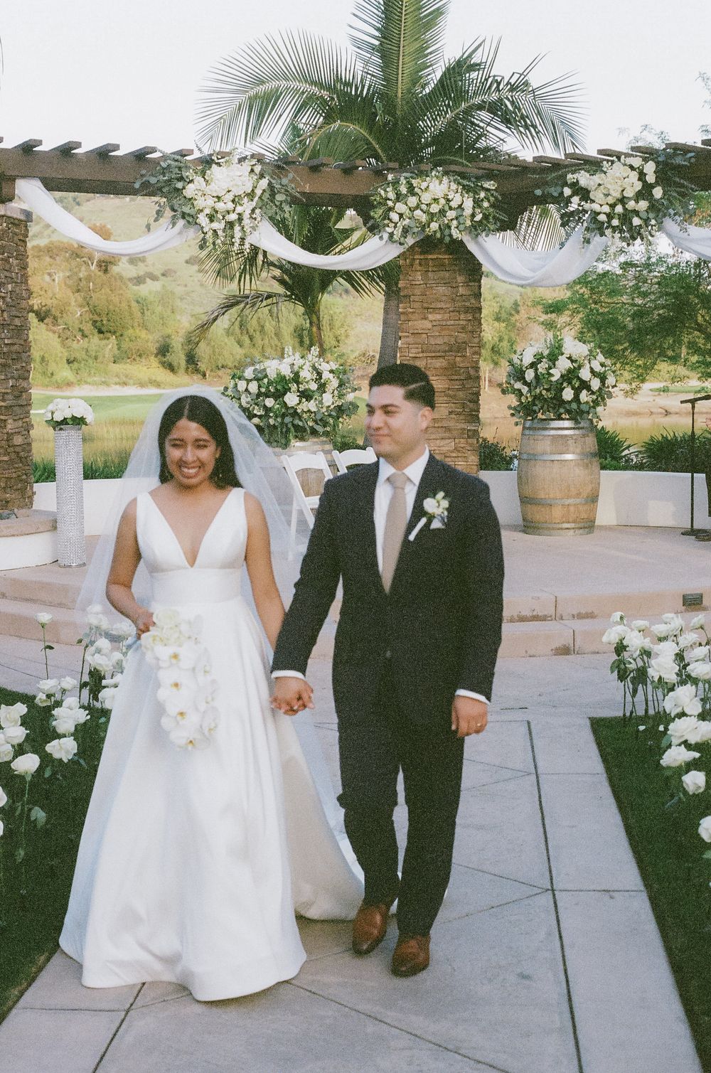 Joyful wedding couple walking hand in hand on stone pathway with tropical garden decor.