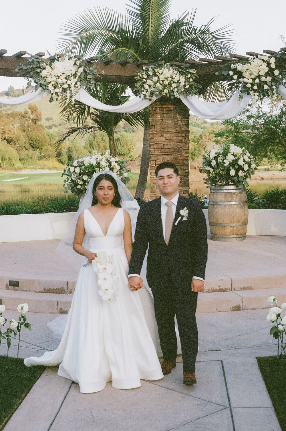 A couple shares a playful moment during their wedding photoshoot in a scenic outdoor venue with palm trees and floral decor.