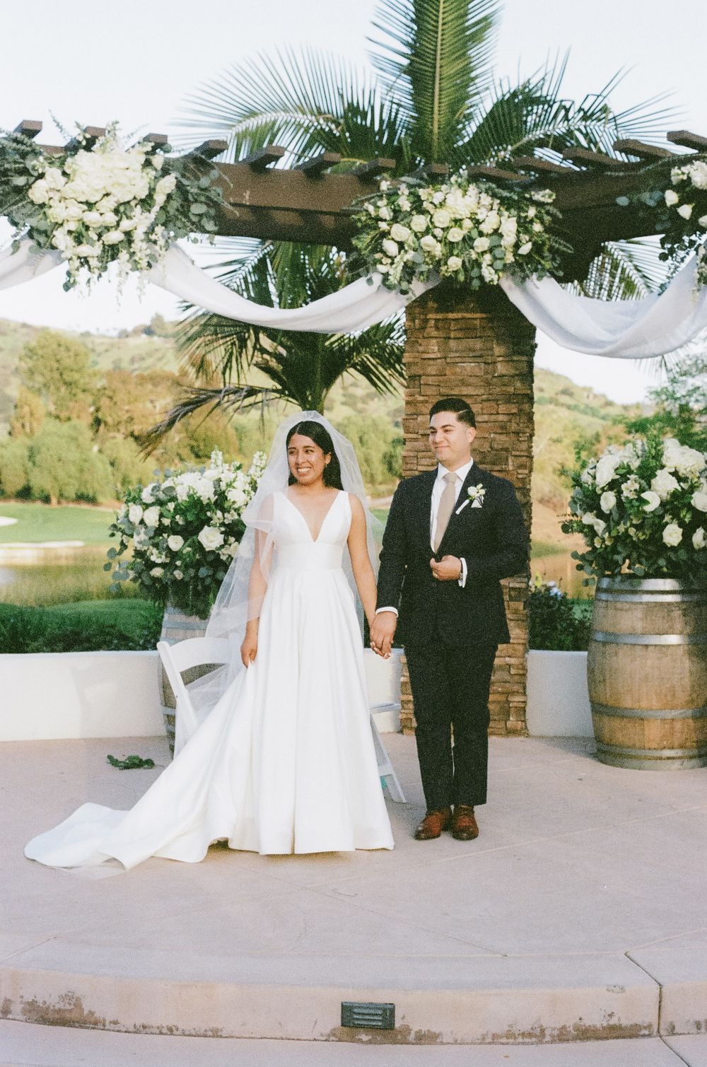 Newlyweds pose for candid photos at their outdoor wedding venue with rustic stone columns and barrel decor.