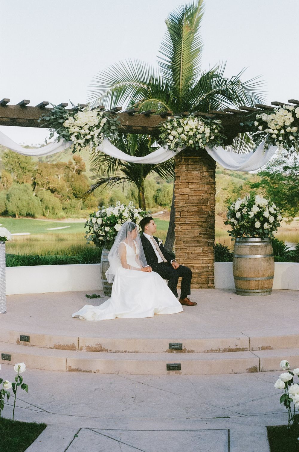 Wedding ceremony venue features stone columns, palm trees and white floral arrangements in an outdoor setting.