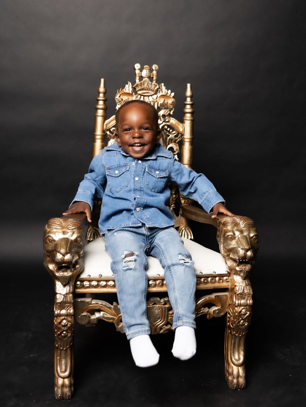 Young child in denim outfit sits smiling on golden throne chair against dark background.