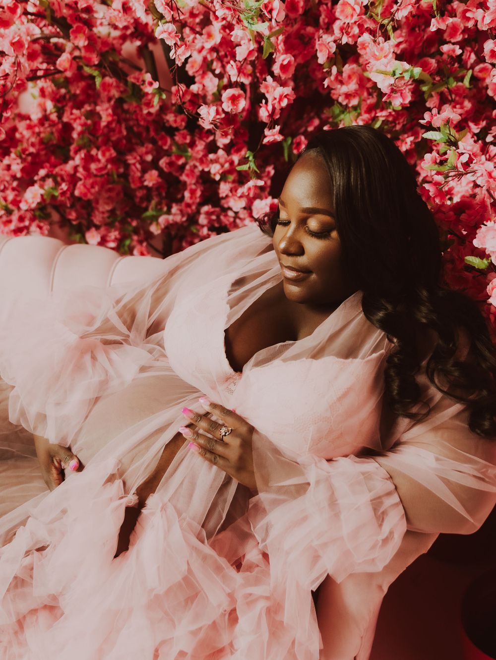 Woman in flowing pink gown poses against floral backdrop for glamour portrait.
