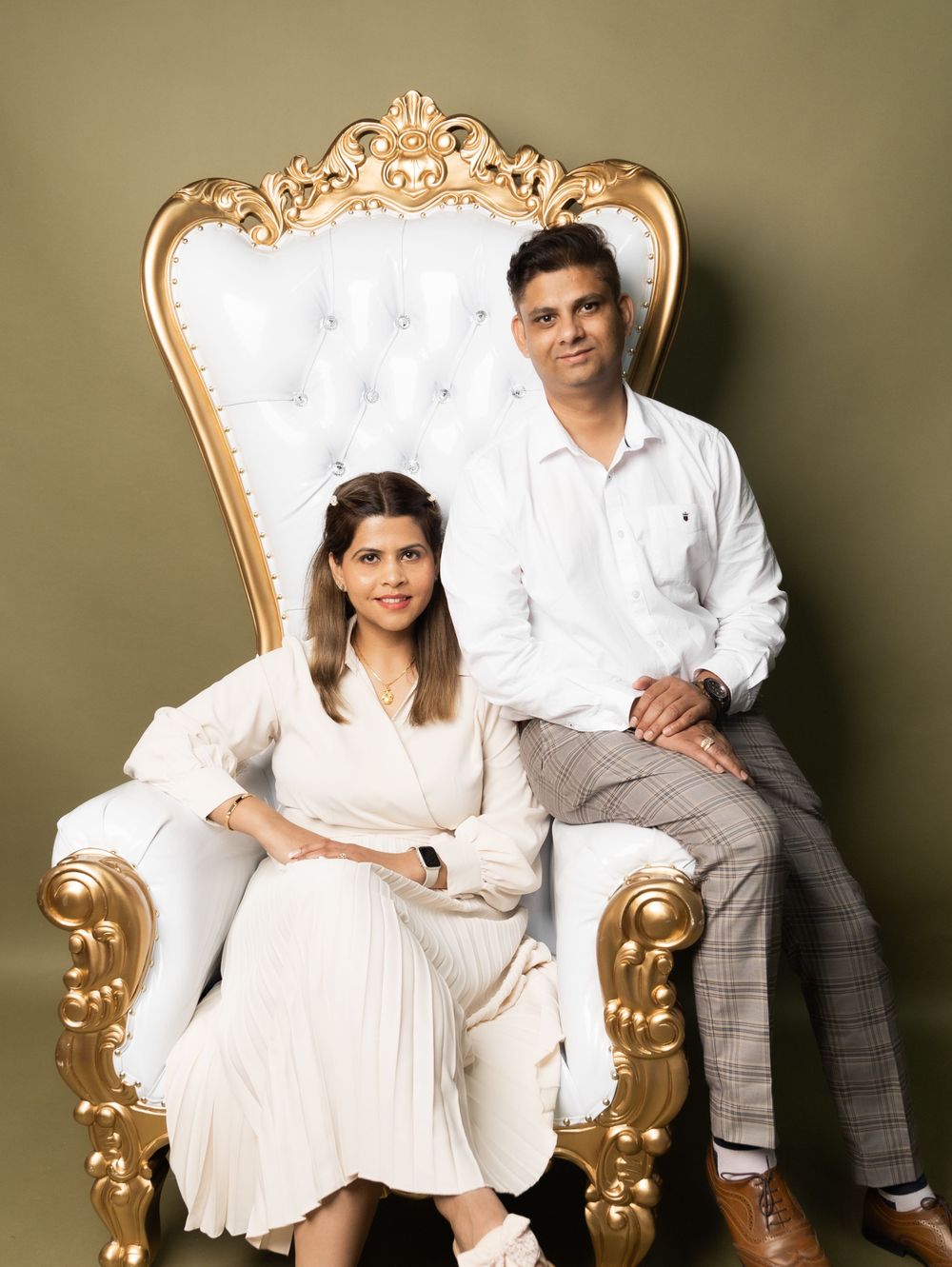 Couple poses on ornate gold and white throne chair for elegant portrait.