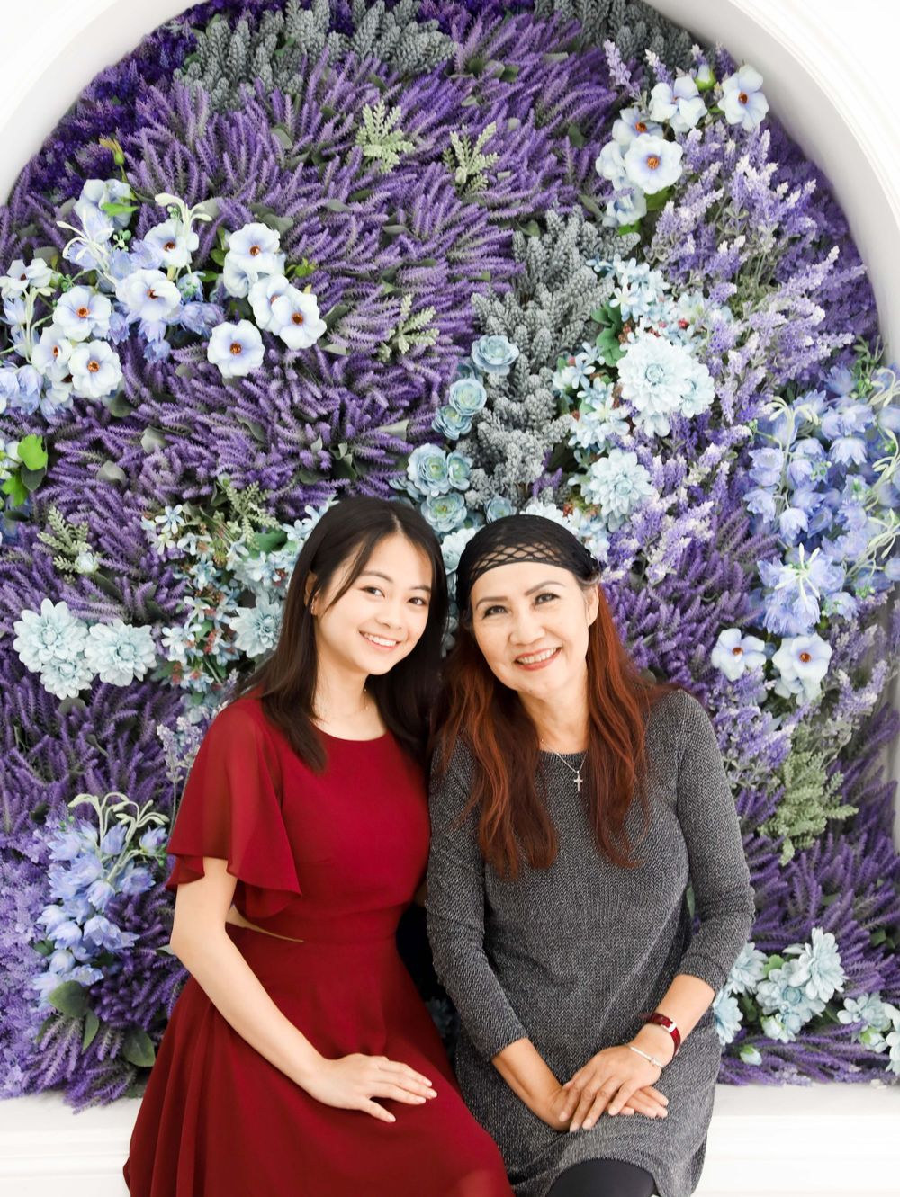 Two women posing against floral backdrop with purple and white blooms.