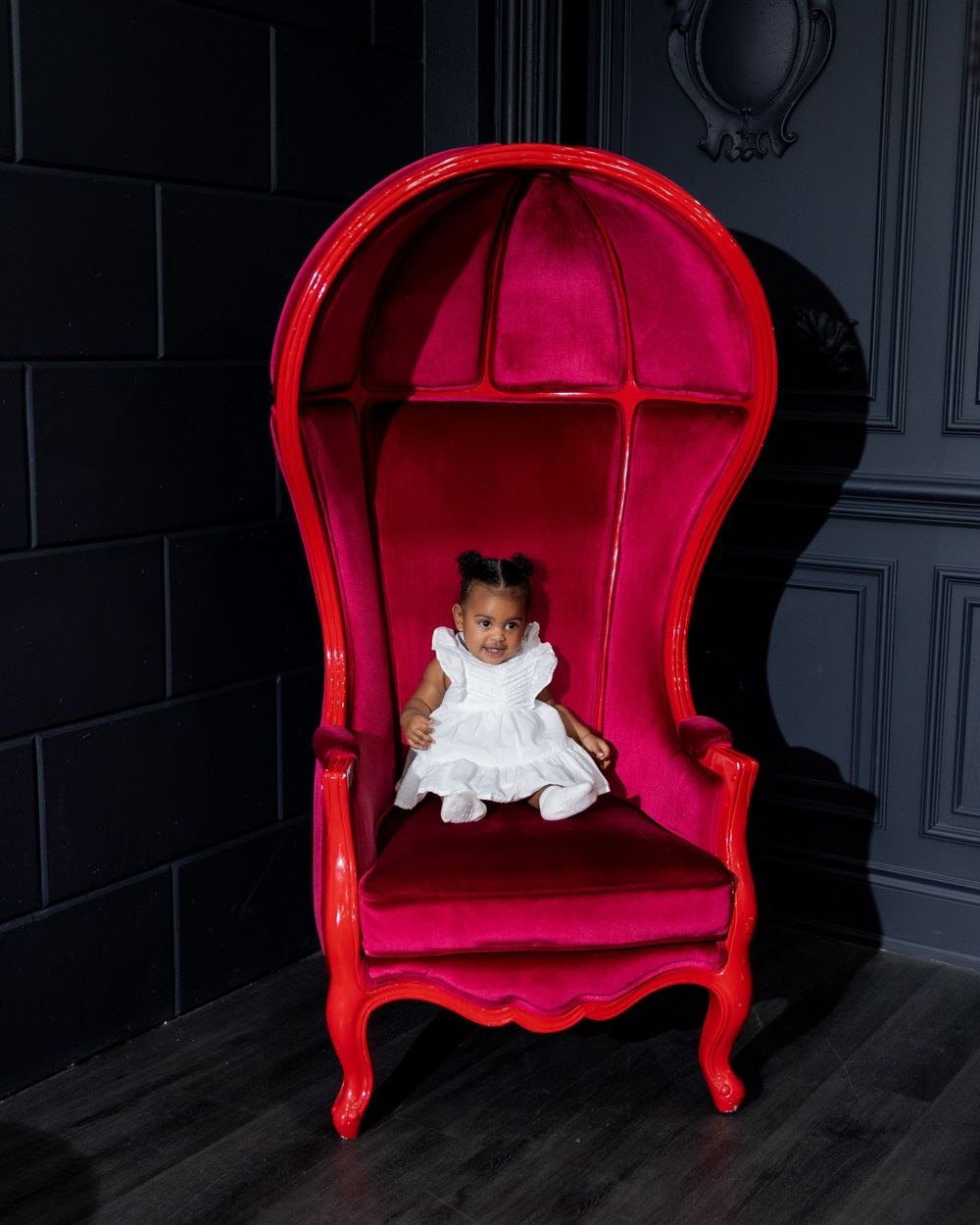 Small child sitting in large red vintage throne chair against dark wall.