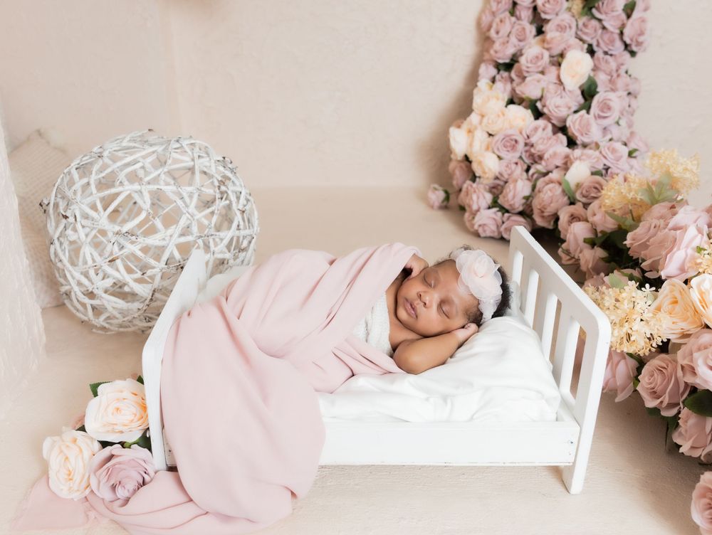 Sleeping newborn wrapped in pink blanket lies in white crib surrounded by flowers.