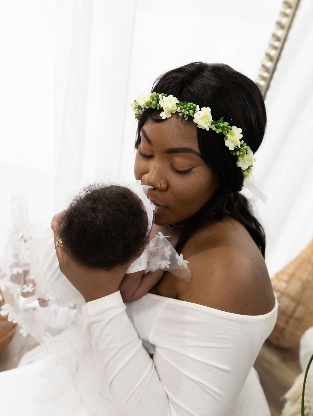 Mother wearing floral crown holds baby in tender moment against white background.