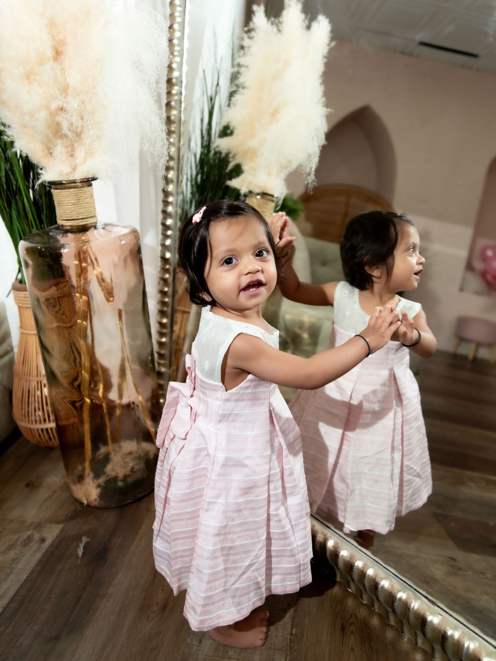 Young child in pink dress smiling at reflection in mirror next to decorative pampas grass.