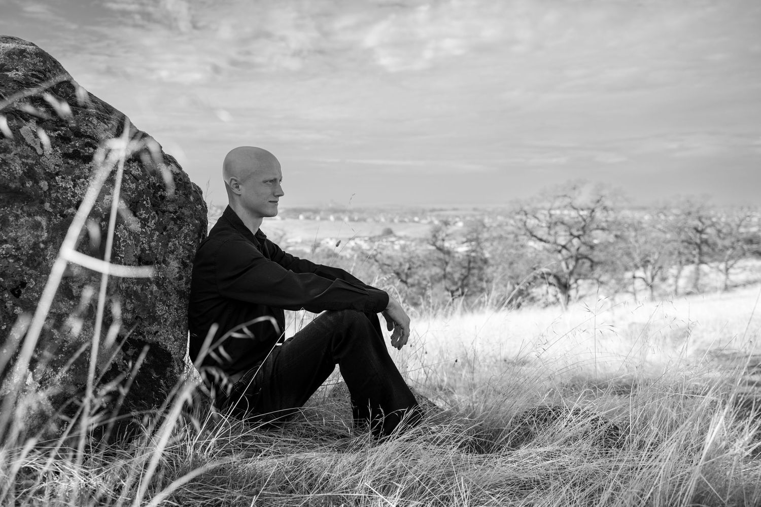Person in dark clothing sits contemplatively on hillside overlooking mountain landscape.
