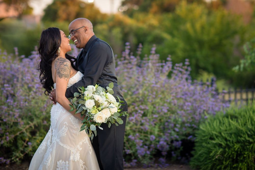 A couple shares a romantic moment in a garden with purple flowers and lush greenery during a wedding photoshoot.
