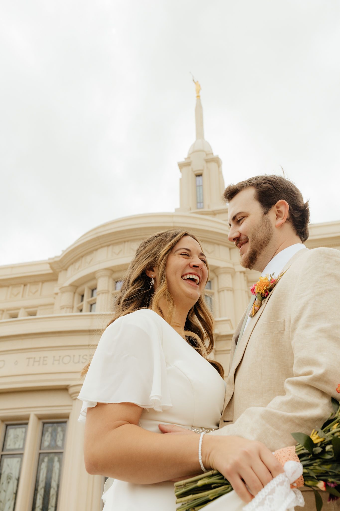 Joyful bride and groom laugh together in front of an LDS temple on their wedding day.