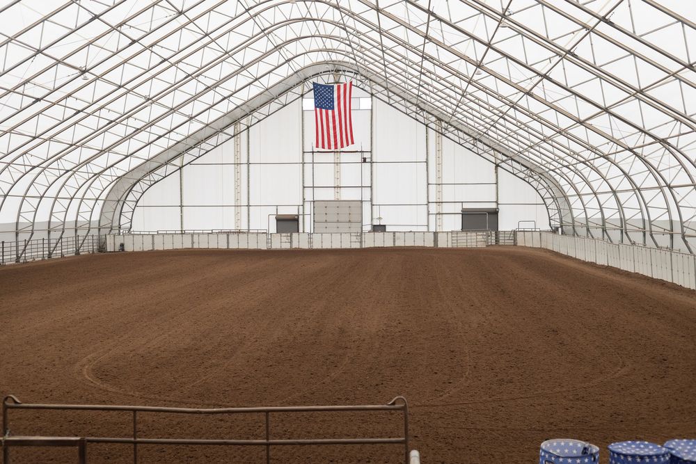 Large indoor equestrian arena with dirt floor, white fabric roof, and American flag displayed on the end wall.