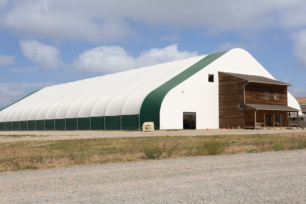 Large white fabric tension structure with green trim beside a wooden building on a rural gravel lot.
