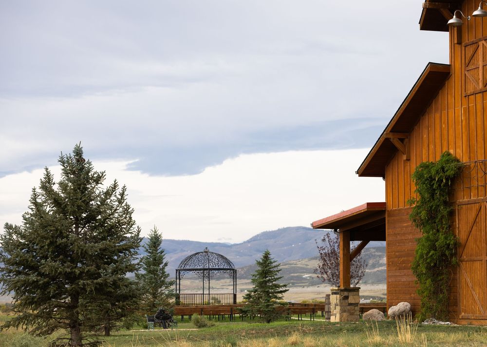Rustic wooden barn with evergreen trees, metal pergola, and mountain views under cloudy skies in a rural setting.