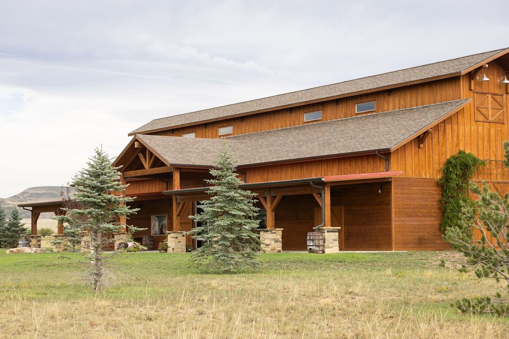 Large rustic wooden barn with stone pillars, evergreen trees, and open fields under a cloudy sky.