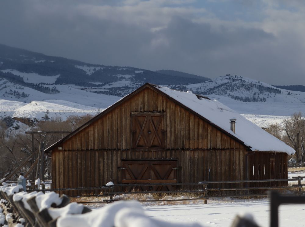 Snow-covered wooden barn set against winter mountain landscape with storm clouds overhead.