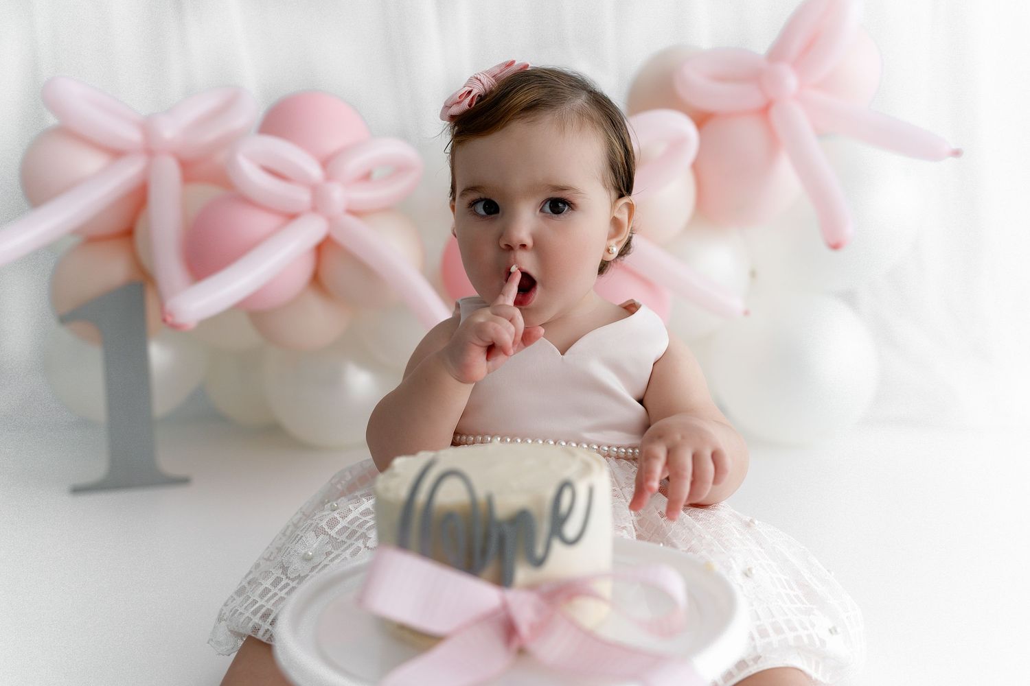 Baby in white dress sits on decorative chair with pink bow balloons in background.