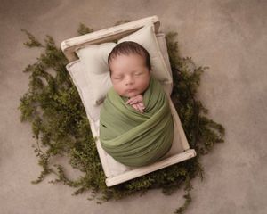 A sleeping newborn wrapped in sage green fabric rests in a wooden box surrounded by moss.