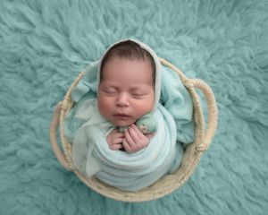 Sleeping newborn swaddled in mint wrap posed in woven basket on teal fur.