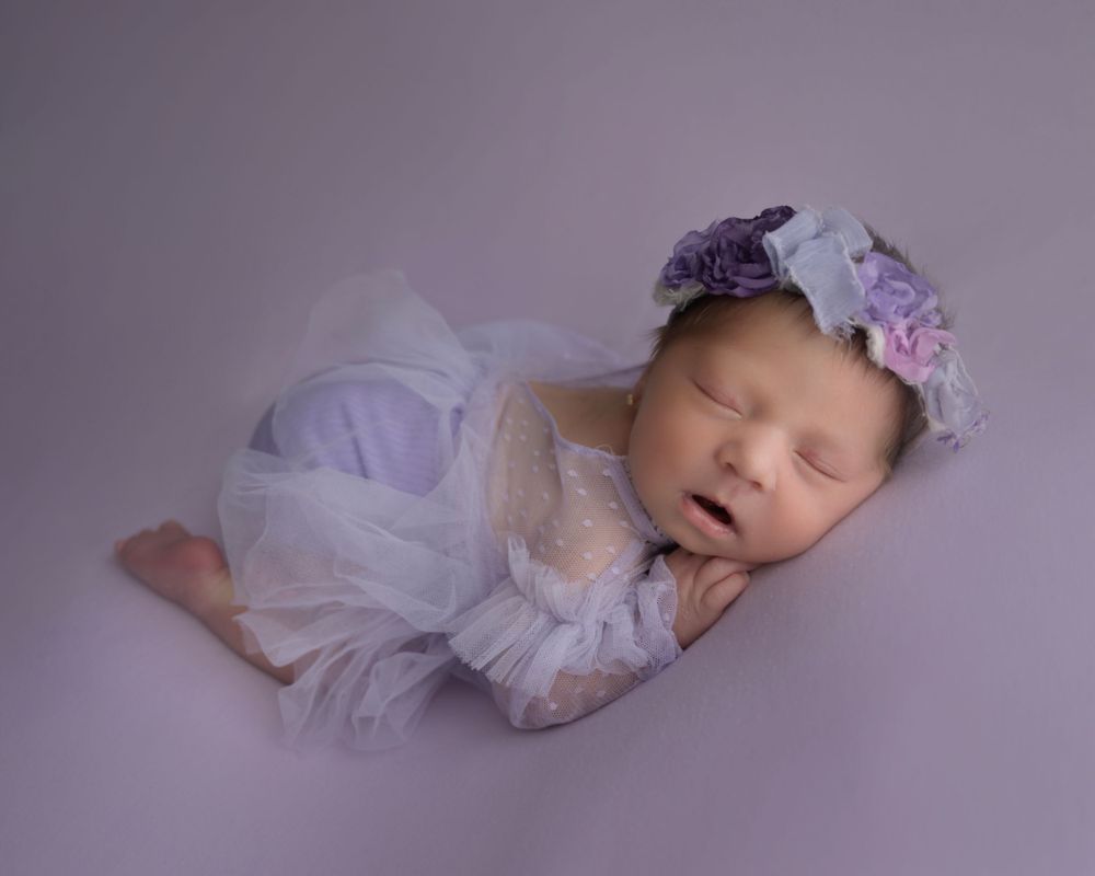 Peaceful newborn wearing white lace dress and purple floral crown on lavender background.
