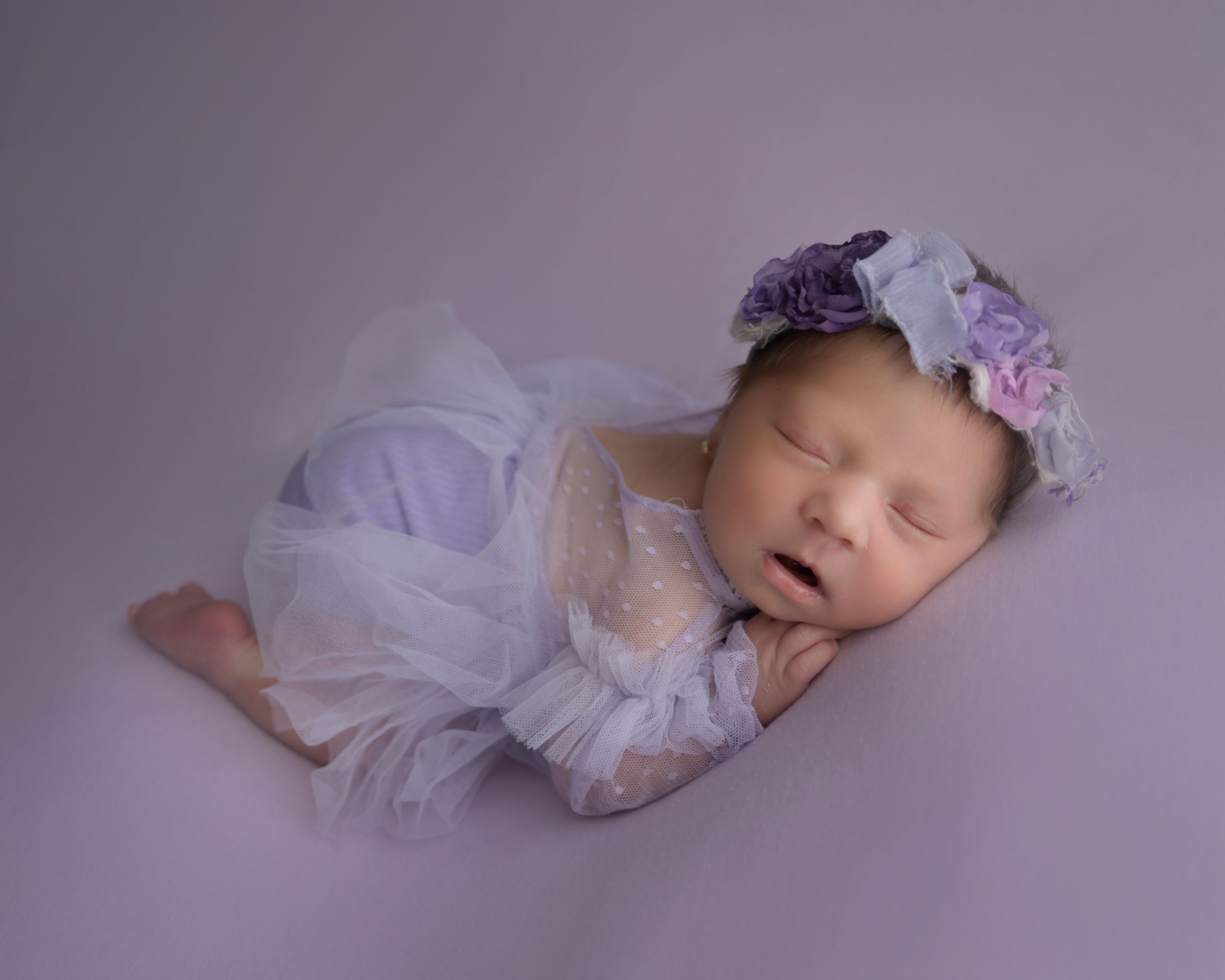 Peaceful newborn wearing white lace dress and purple floral crown on lavender background.