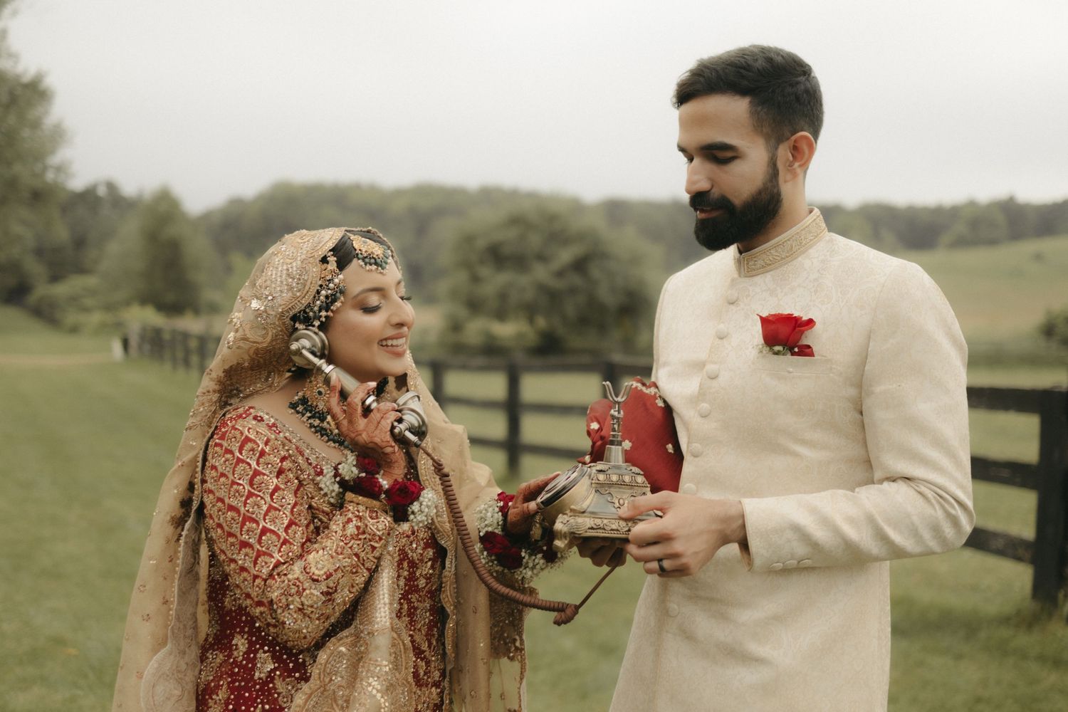 A couple in traditional formal attire sharing a moment in front of a countryside fence.