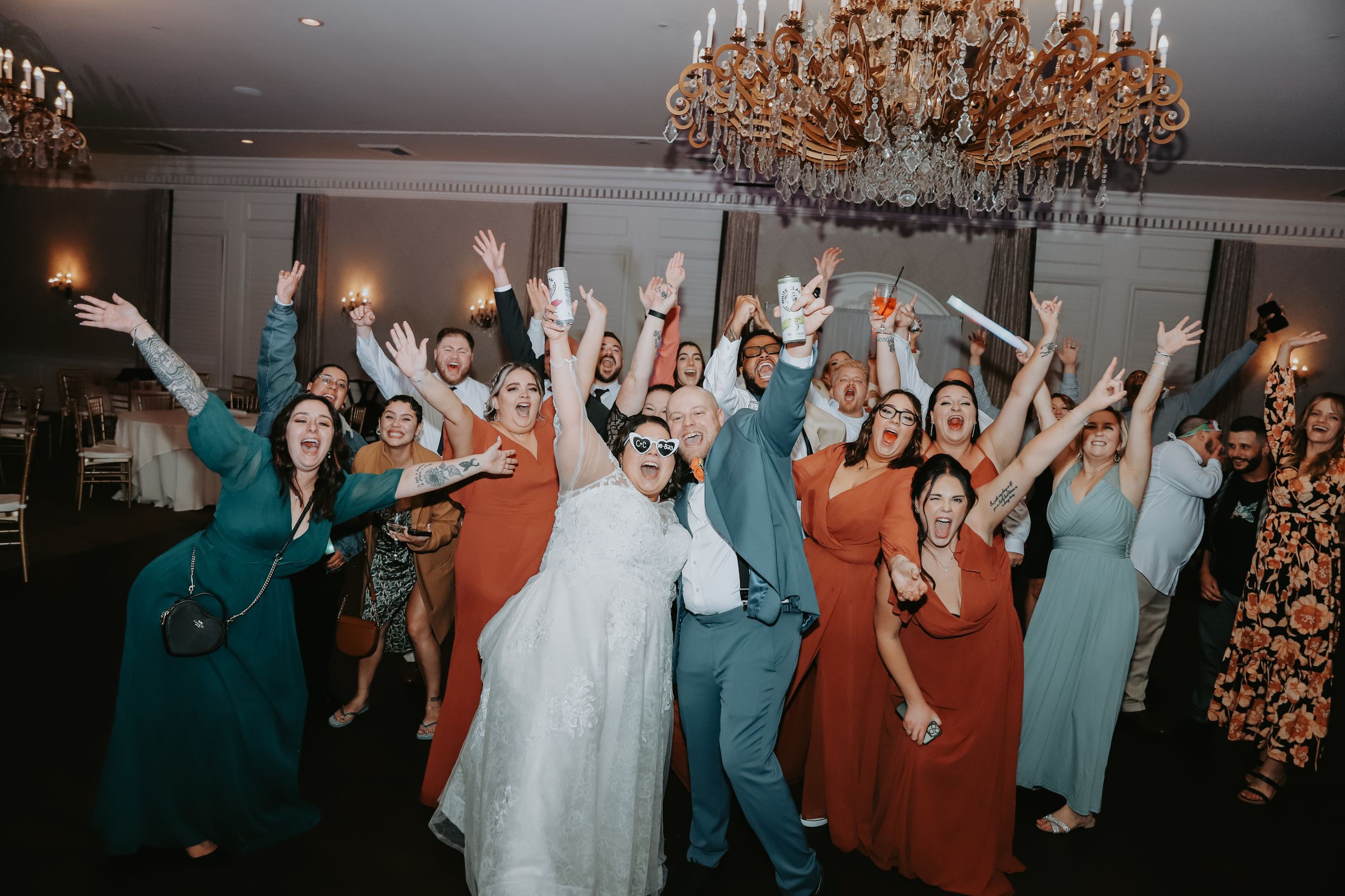 Energetic wedding reception dance party with guests in formal attire under crystal chandelier.