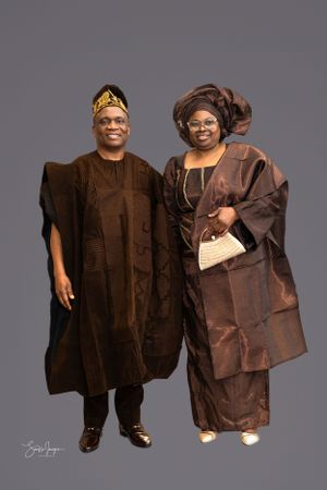Two people wearing traditional brown Nigerian attire including agbada and gele pose together against a gray backdrop.