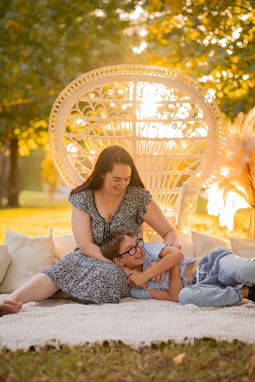 Mother and son laughing together on blanket in front of decorative white peacock chair at golden sunset.