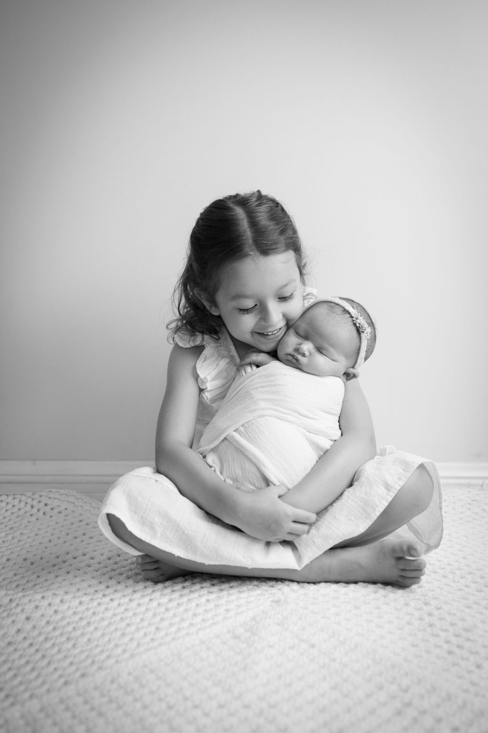 Black and white photo of young girl tenderly holding swaddled newborn baby while seated on floor.