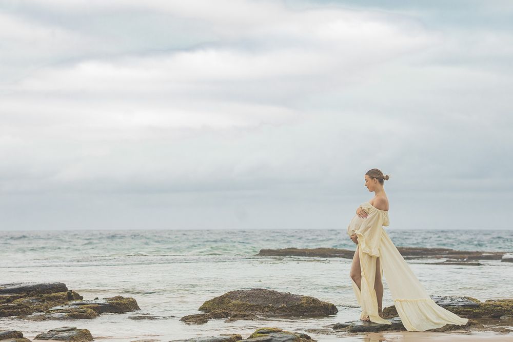 Pregnant woman in elegant white off-shoulder gown standing on rocky beach under cloudy sky.
