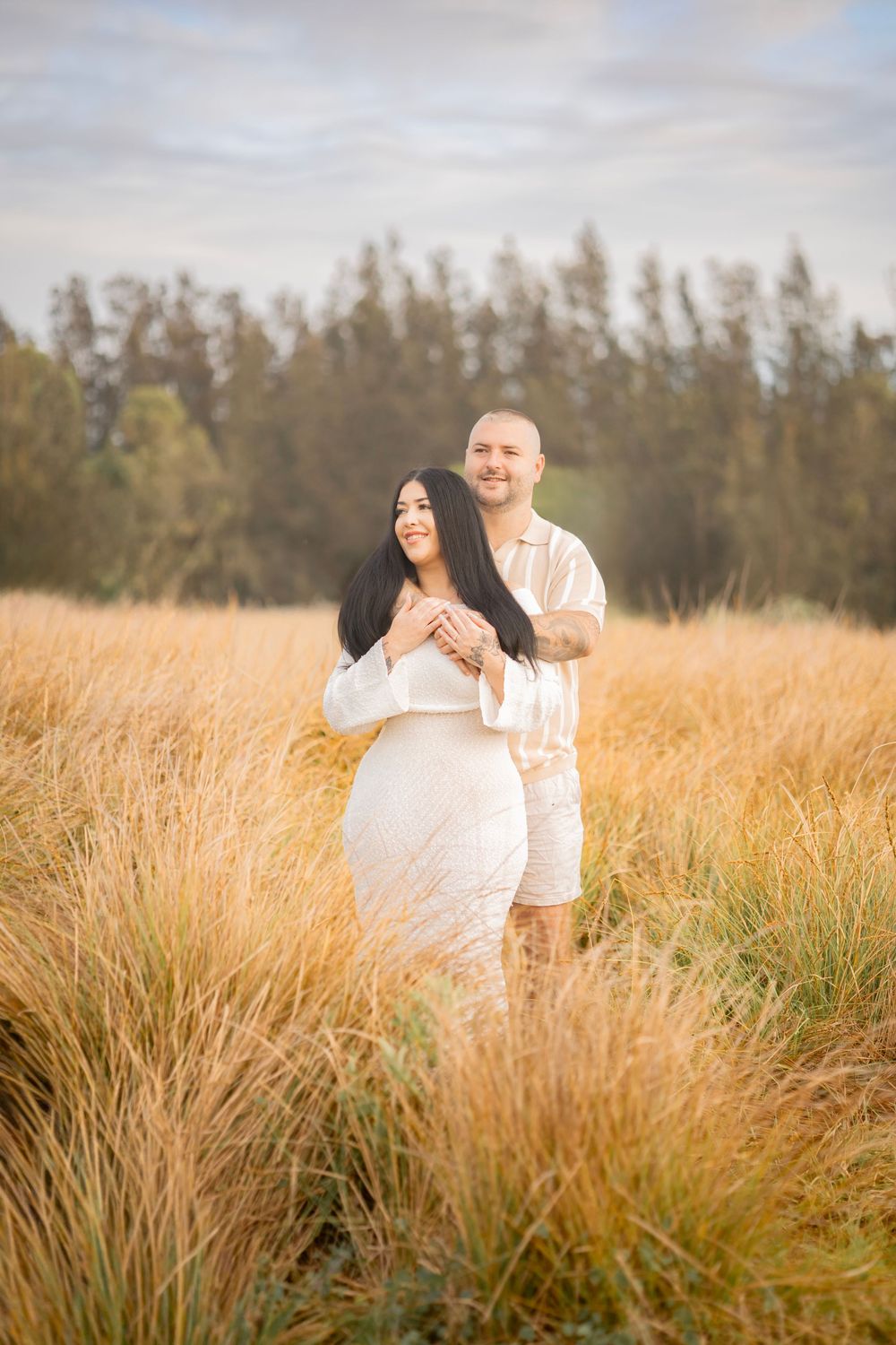 Couple in white outfits embracing in golden tall grass field during soft sunset light.