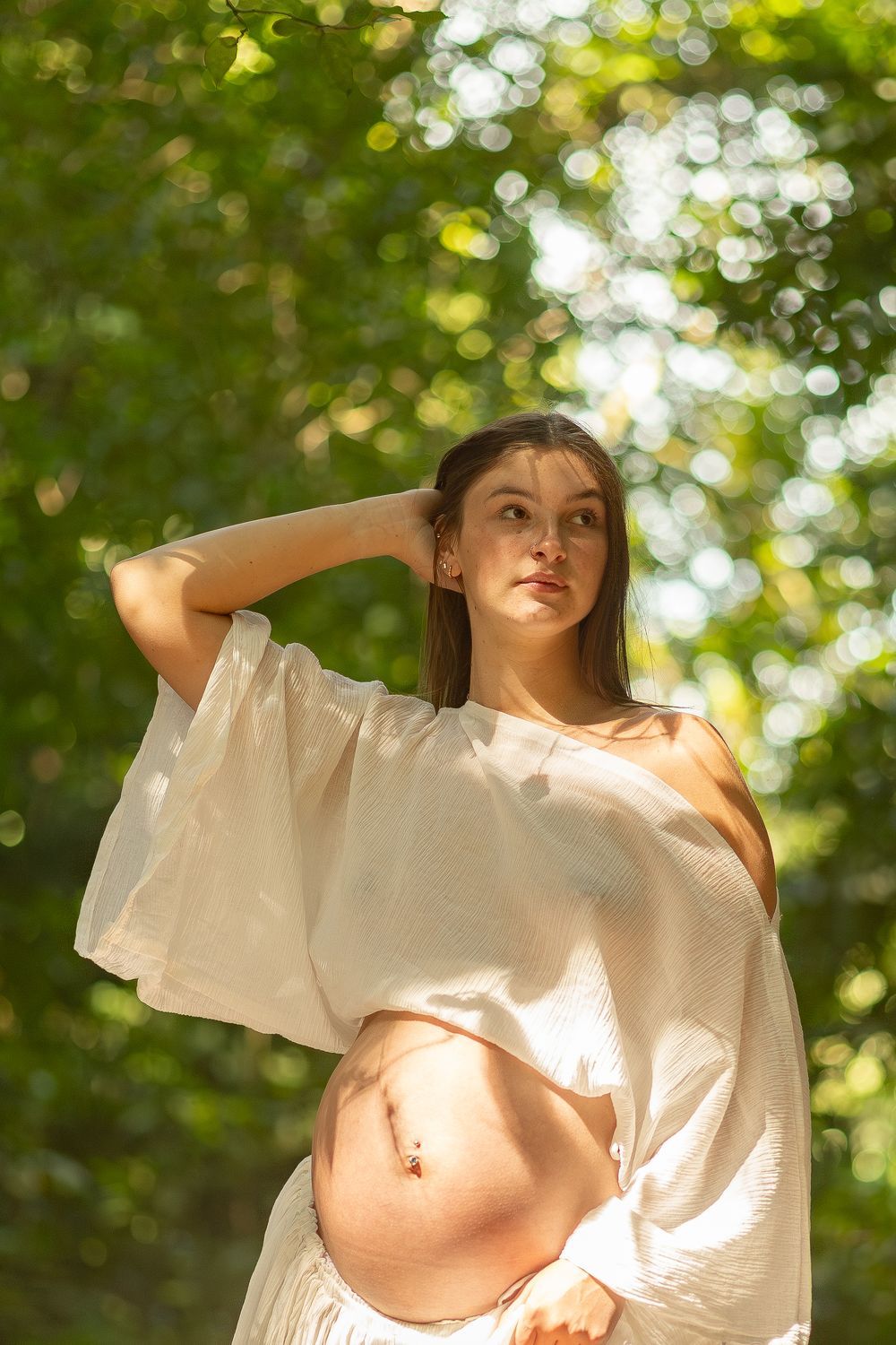 Young pregnant woman in white top posing outdoors in dappled sunlight with green bokeh background.