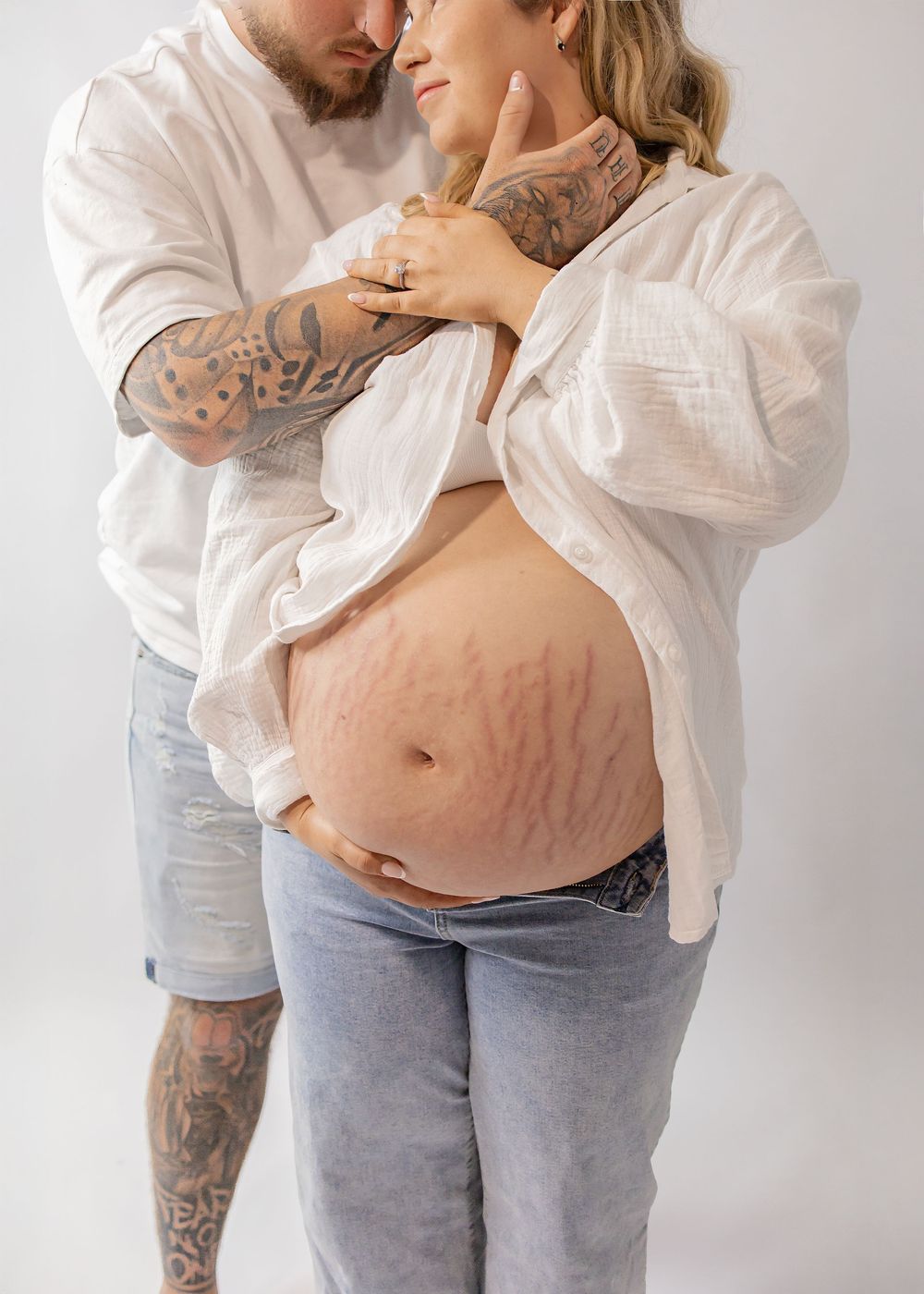 Tattooed couple in white outfits embracing while proudly displaying pregnant belly with stretch marks.