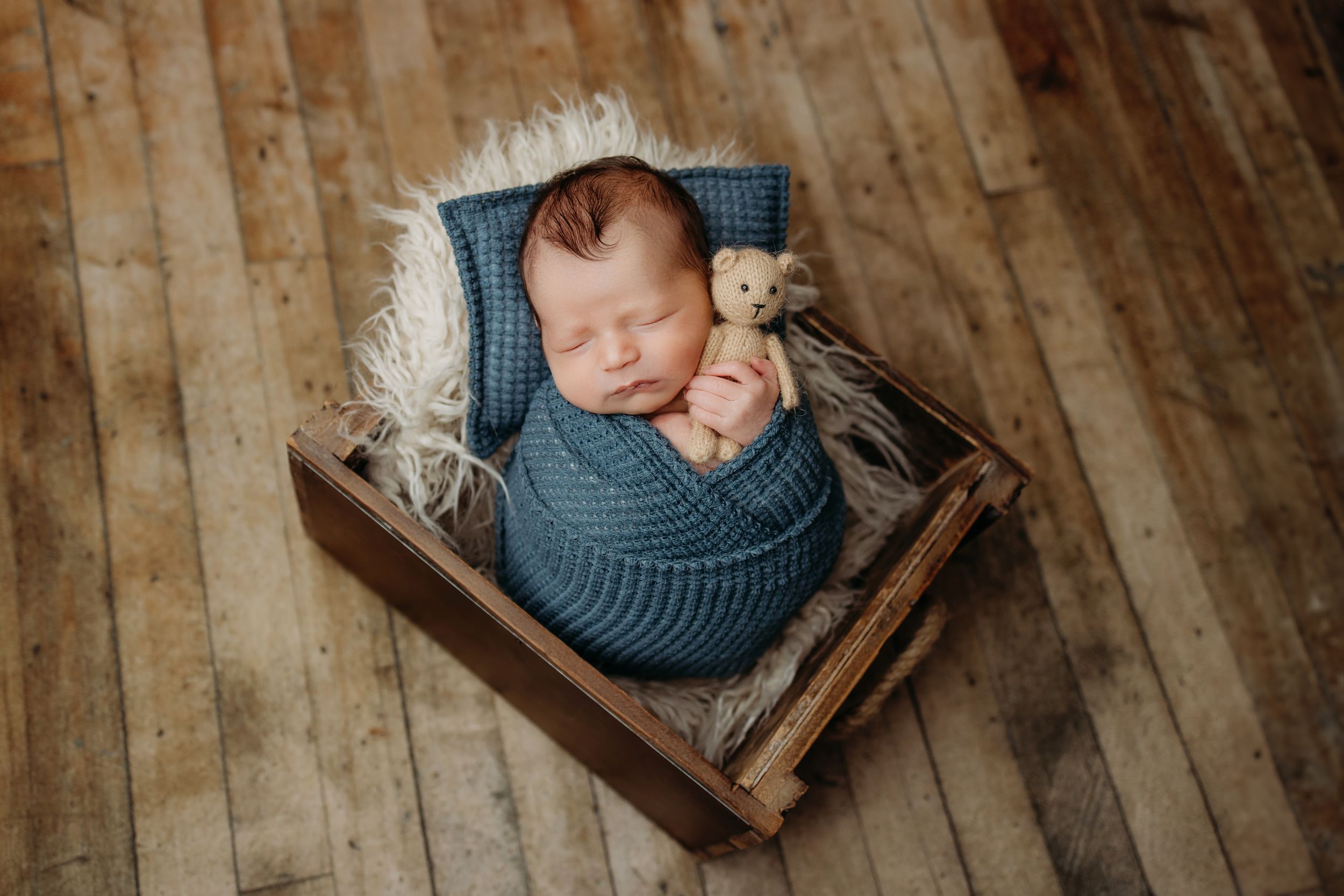 A sleeping newborn wearing blue swaddle cloth rests peacefully in a wooden crate during a photography session.