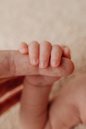 A tiny baby's hand grasps an adult's finger in a tender close-up shot against a soft beige background.