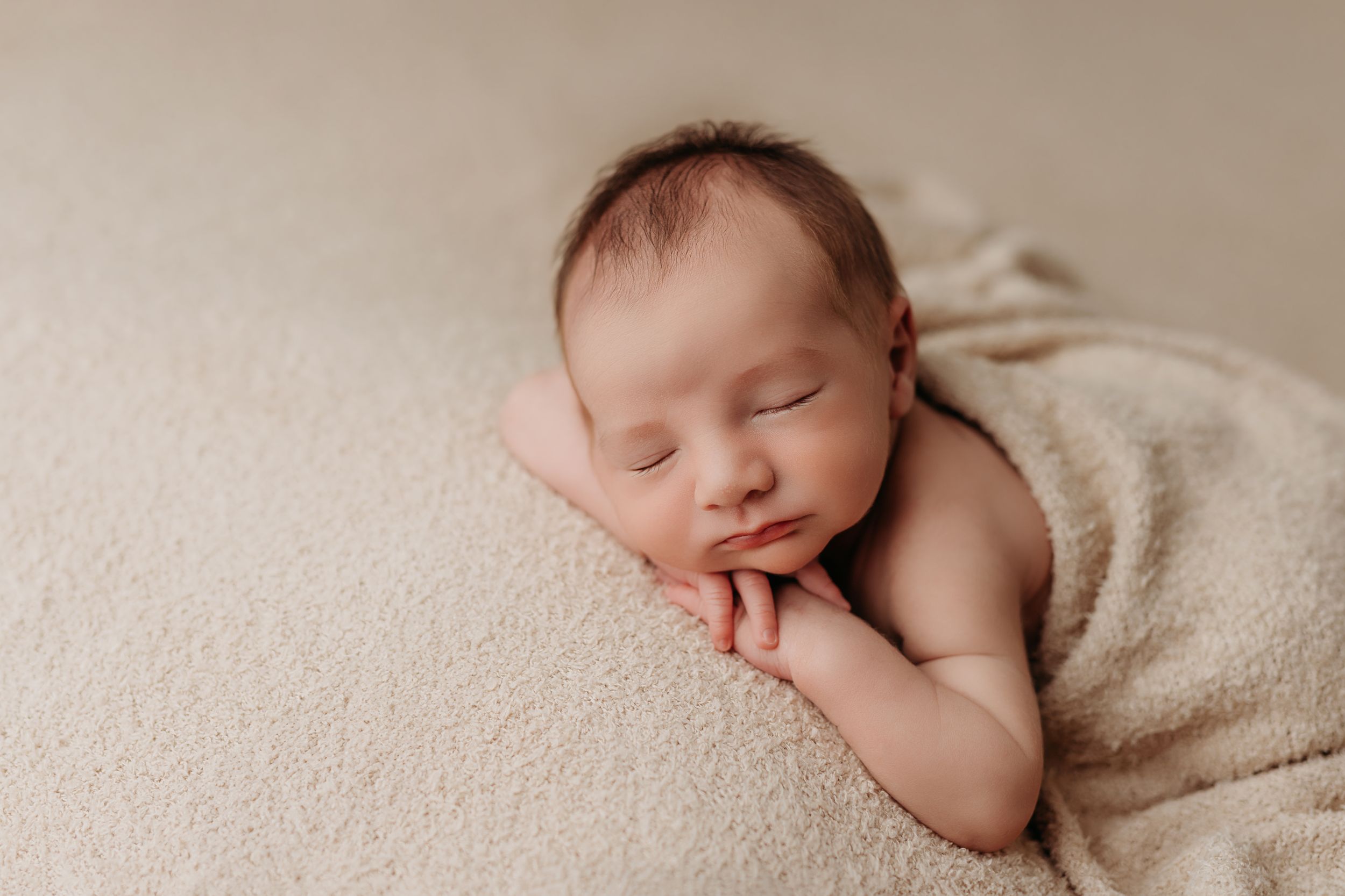 A peaceful sleeping newborn rests on a cream-colored blanket with one hand tucked under their cheek.
