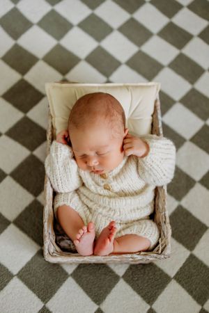 Newborn baby sleeping peacefully in white knit outfit on checkered floor pattern.