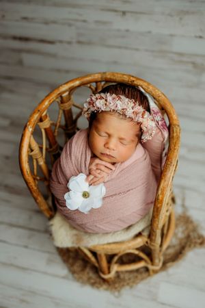 Sleeping newborn in pink wrap with floral headpiece resting in bamboo chair.