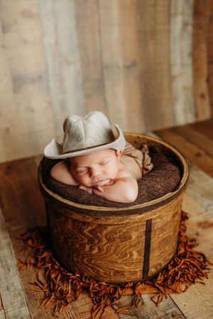 Sleeping newborn in grey hat resting in wooden bucket on a wooden backdrop.