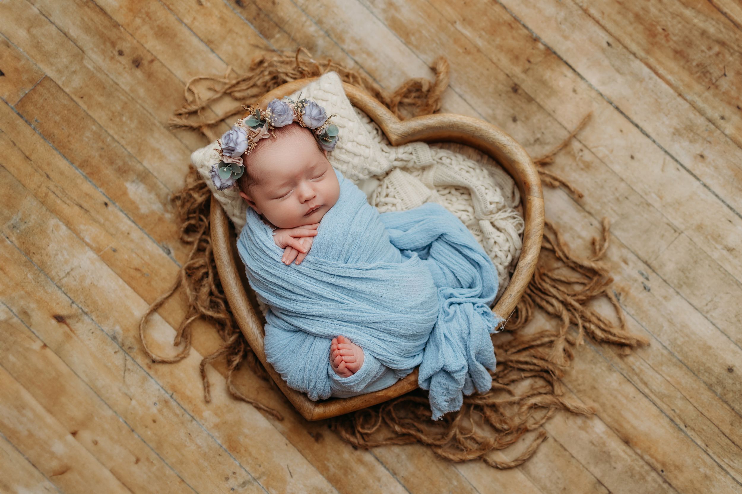 Newborn baby wrapped in light blue swaddle blanket lying on rustic wooden floor in professional photography setup.
