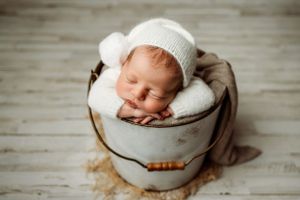 Peaceful newborn wearing white knit hat nestled in cozy white bucket.