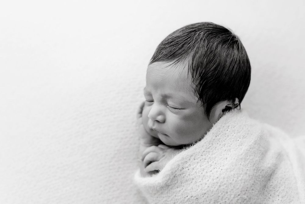 A sleeping newborn baby rests peacefully on soft white fabric in black and white.