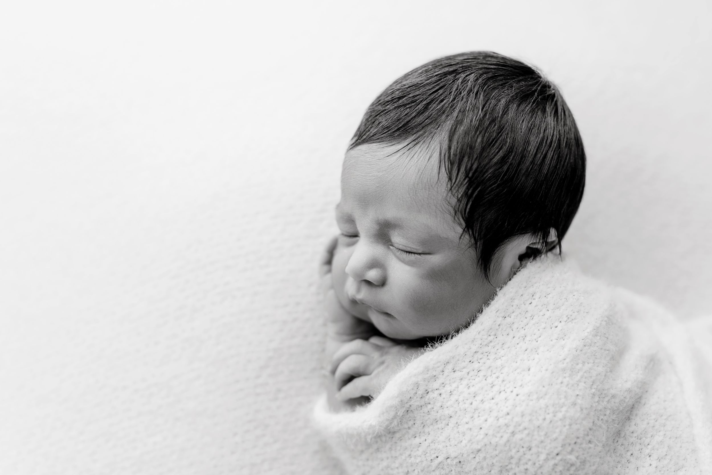 A sleeping newborn baby rests peacefully on soft white fabric in black and white.
